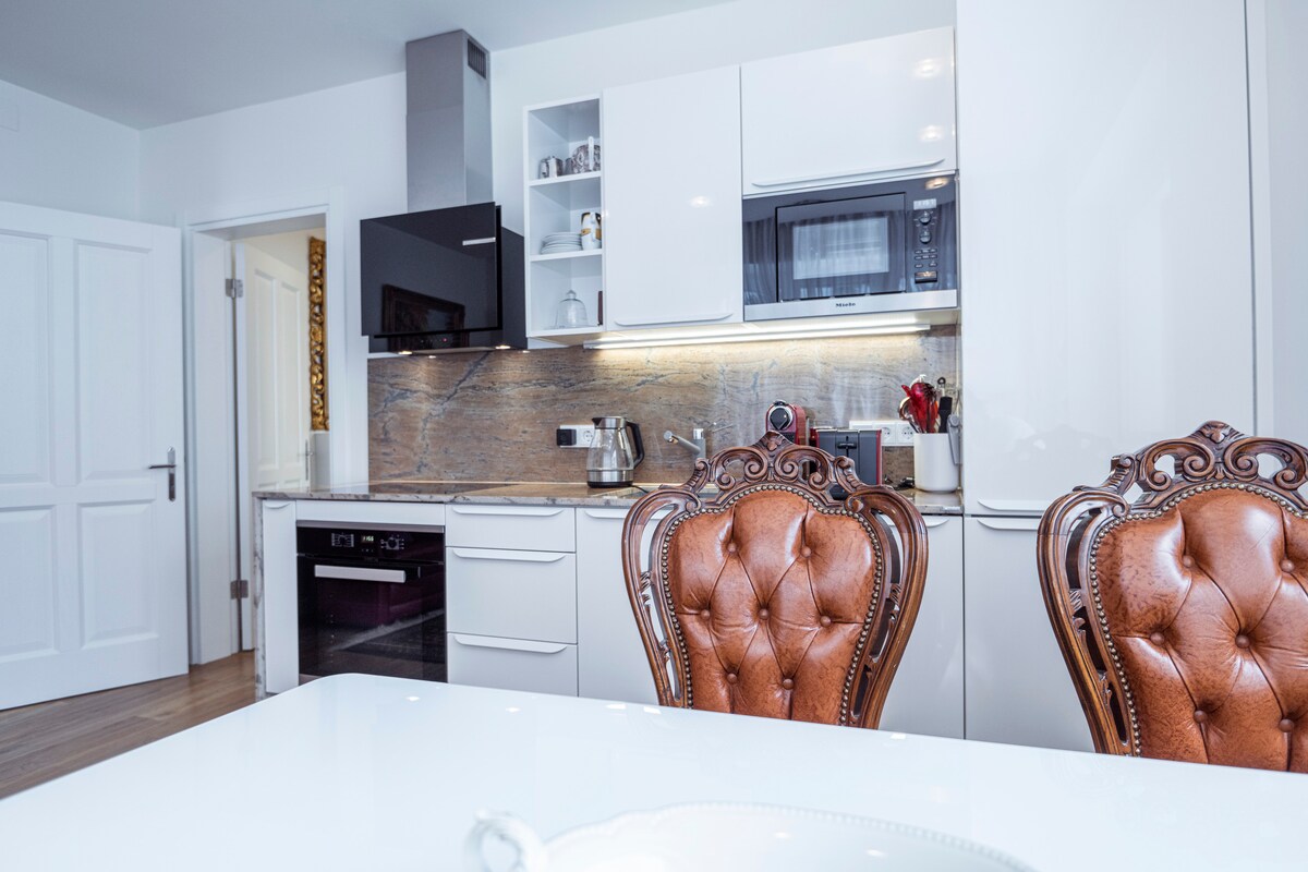 The kitchen area features modern cabinetry with a sleek design, complemented by a stylish countertop. A microwave and oven are integrated within the cabinetry. Two ornate chairs with rich, brown upholstery provide seating around a polished dining table.