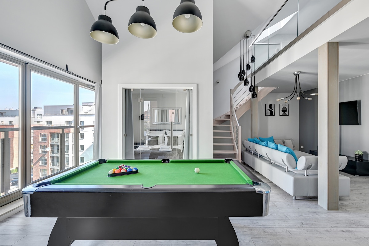 A spacious game area features a billiard table with a bright green felt surface, complemented by pendant lighting above. Large windows allow natural light to fill the room, revealing a modern living space adorned with white furnishings and vibrant blue accents in the background.
