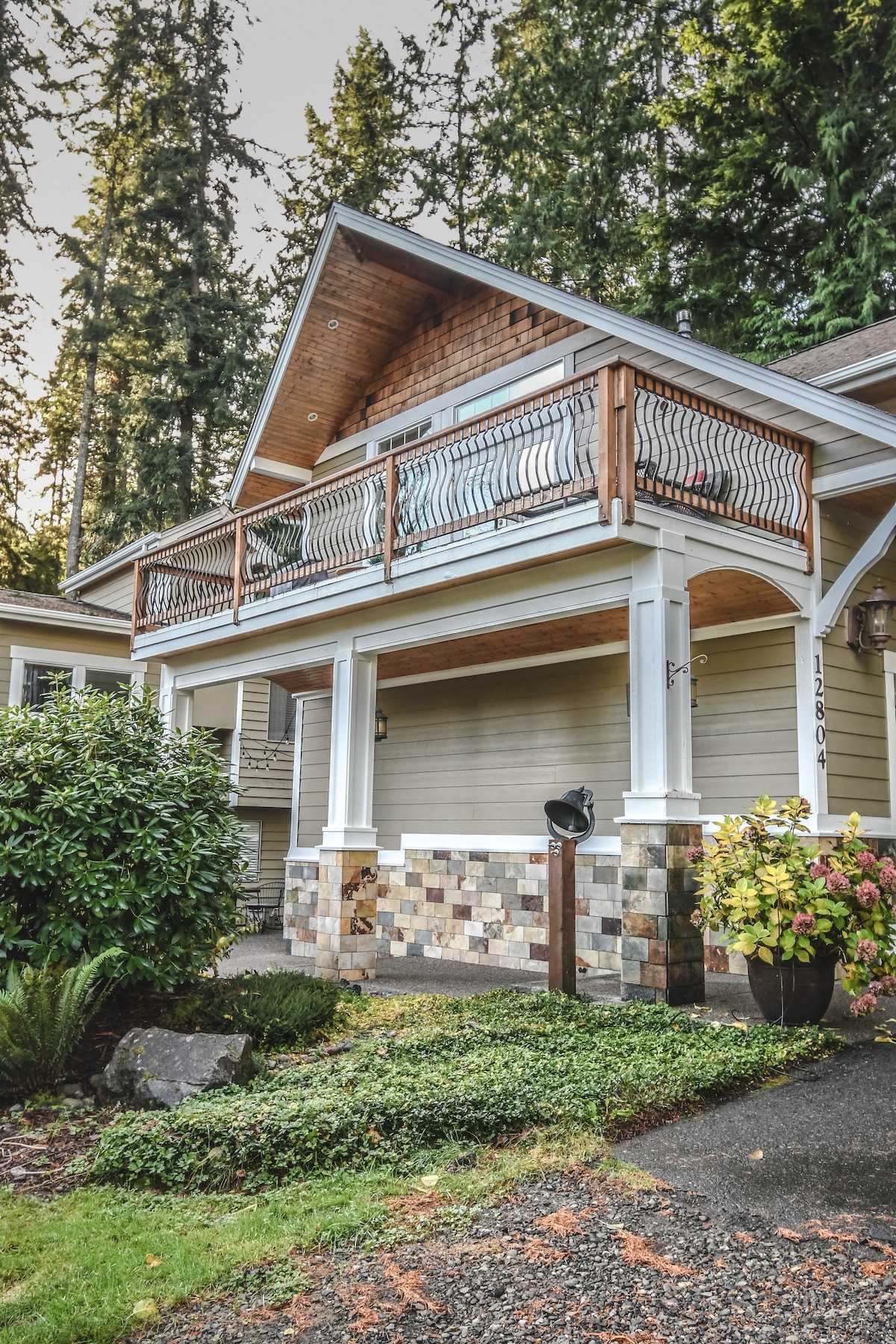 The exterior of the loft is showcased, featuring a welcoming entrance with a stone-accented lower facade. A wooden balcony is supported by ornate railings, surrounded by tall trees, and lush greenery. A pathway leads to the entrance, adding to the inviting appearance.