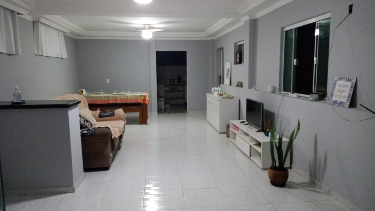 A spacious living area features tile flooring and light gray walls. A soft sofa is positioned beside a wooden dining table, and a television is placed on a low white unit. Large windows provide natural light, and a potted plant adds a touch of greenery.