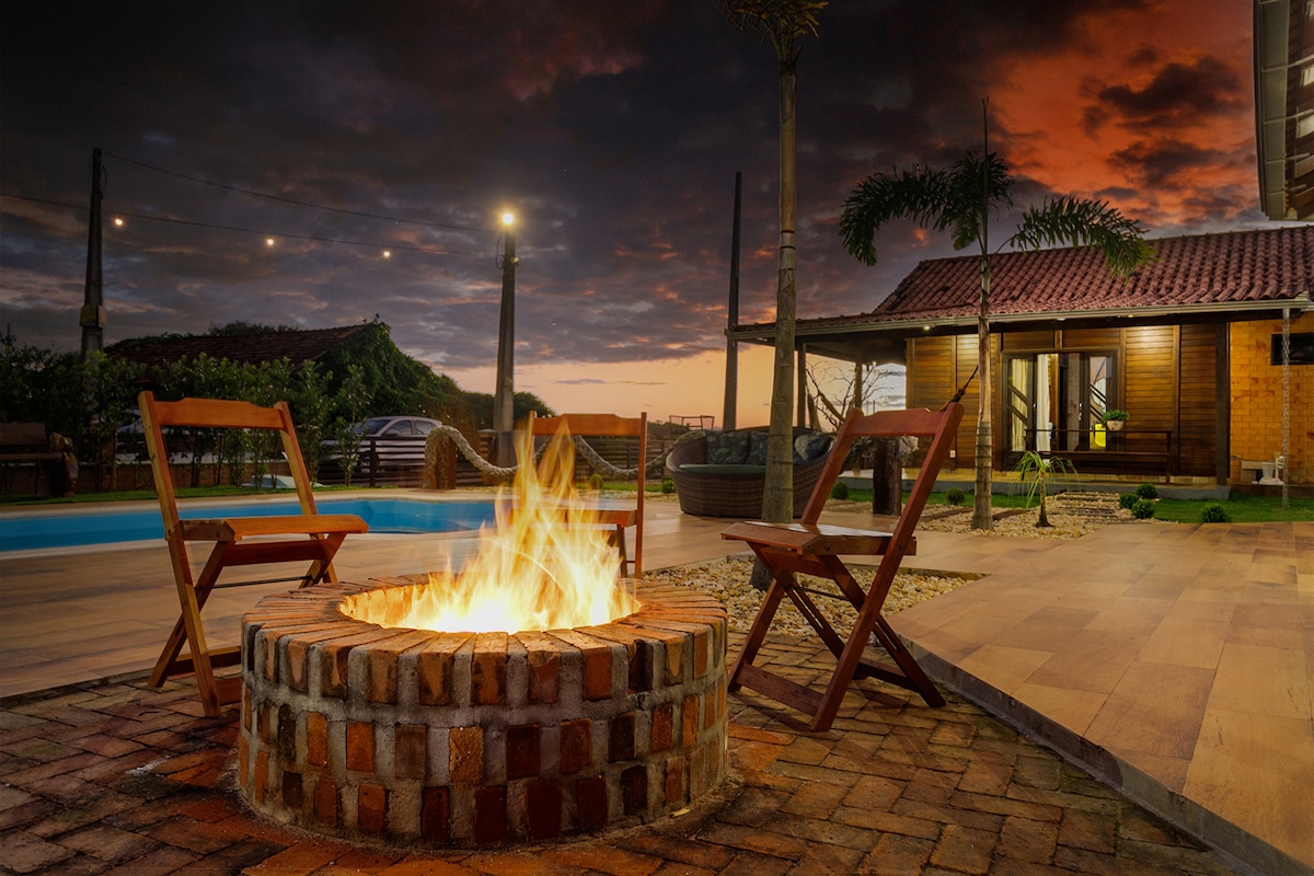 A circular brick fire pit is surrounded by two wooden chairs, inviting gatherings under a dramatic sunset sky. In the background, a tropical landscape and a well-lit house are visible, complementing the serene outdoor atmosphere.