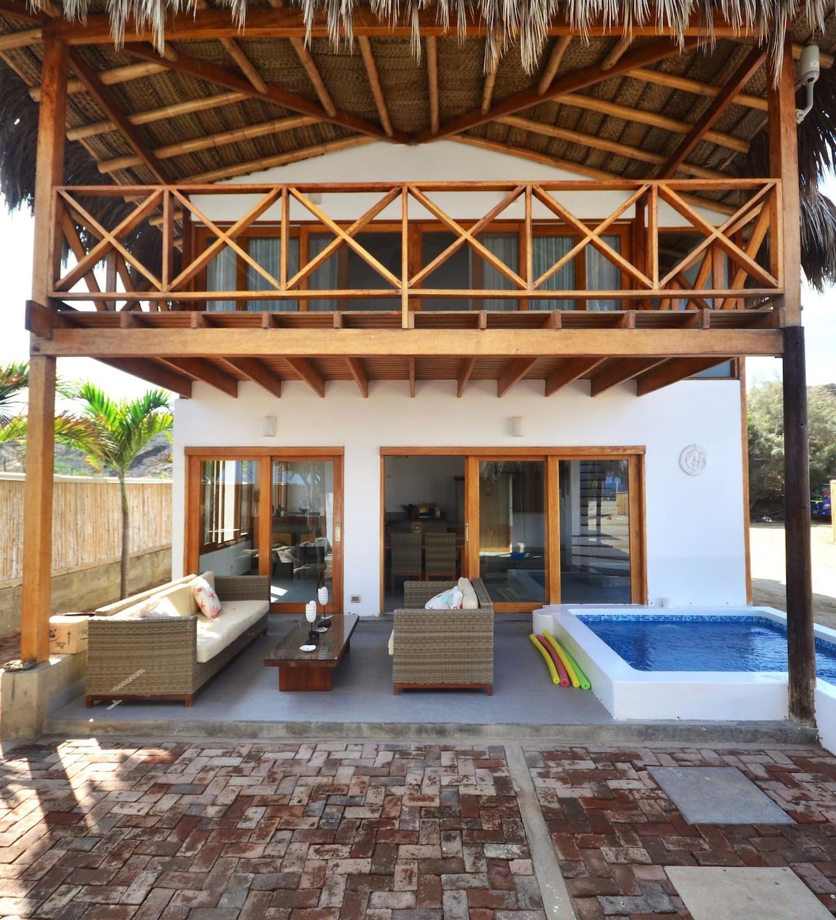 The two-story house is showcased from the front, featuring a thatched roof and large glass doors that open to the outdoor space. A small private pool is visible beside a seating area with cushions. Lush greenery is seen in the background.