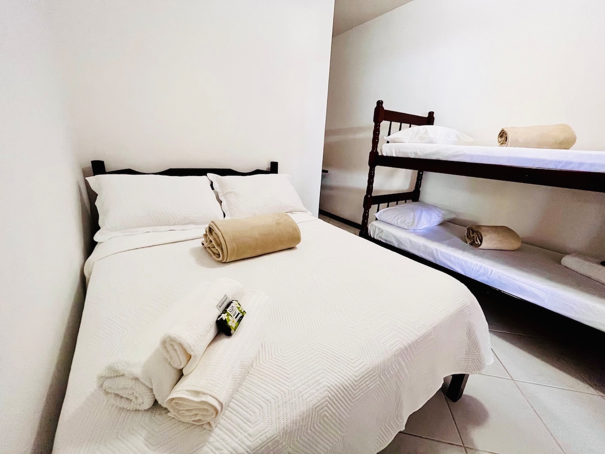 A cozy bedroom features a spacious bed with light bedding, complemented by neatly arranged towels. A set of bunk beds is positioned against the wall, providing additional sleeping space. Soft lighting enhances the room's cleanliness and simplicity.