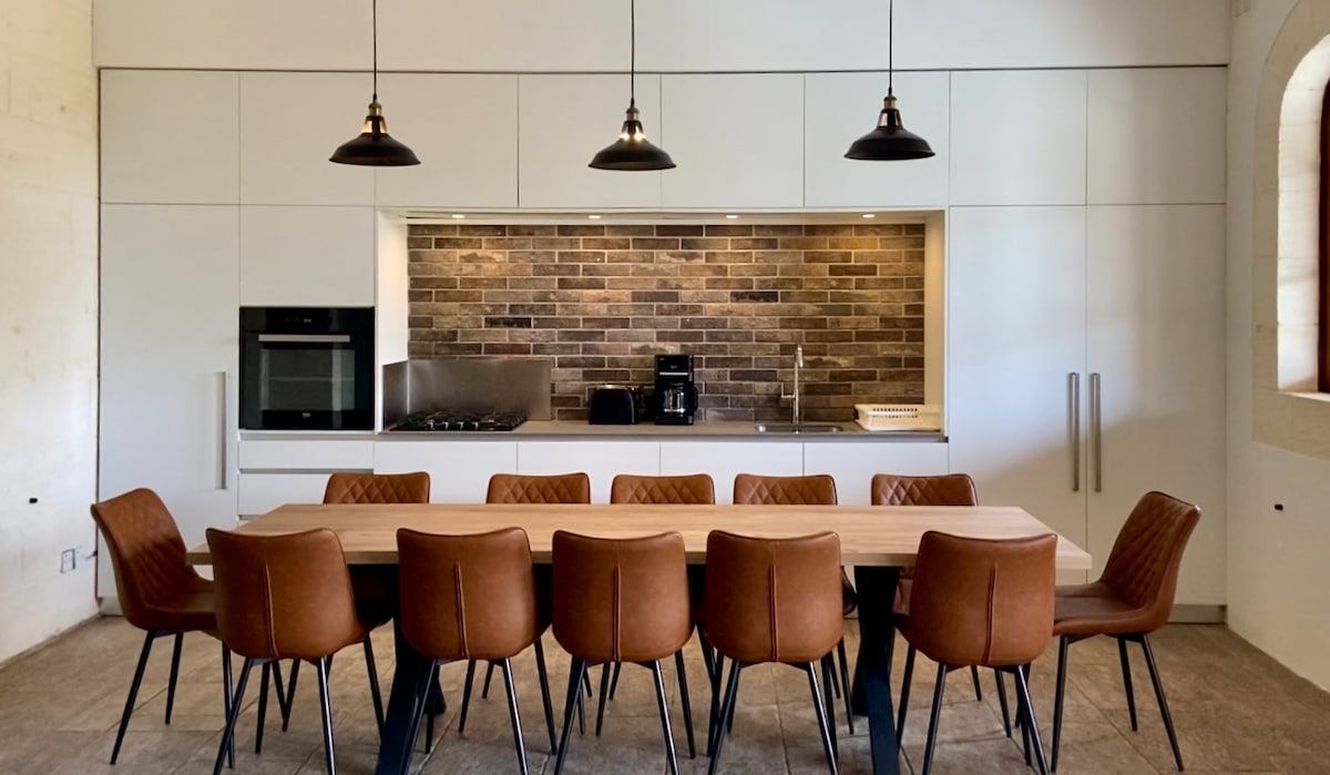 A spacious kitchen area showcases a large wooden dining table with eight brown upholstered chairs. The kitchen features sleek cabinetry, a modern oven, and a stovetop, all complemented by a stylish backsplash of textured bricks. Three pendant lights provide ambient illumination.