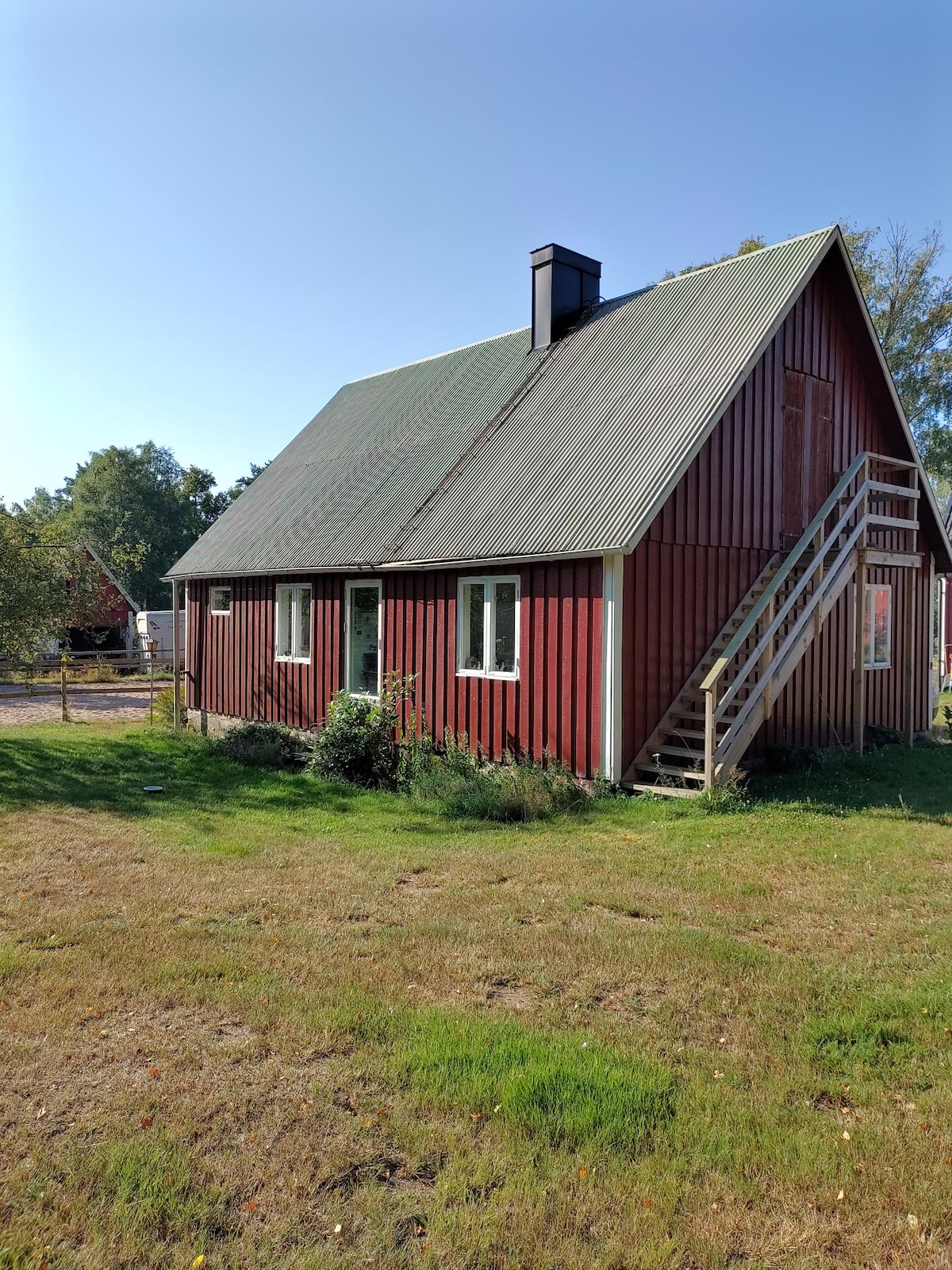 A quaint cottage with a steeply pitched roof is set in a grassy area. The exterior is clad in red wood, complemented by white-trimmed windows. A wooden staircase leads to the entrance. Verdant shrubs and a patch of grass surround the structure, fostering a peaceful setting.