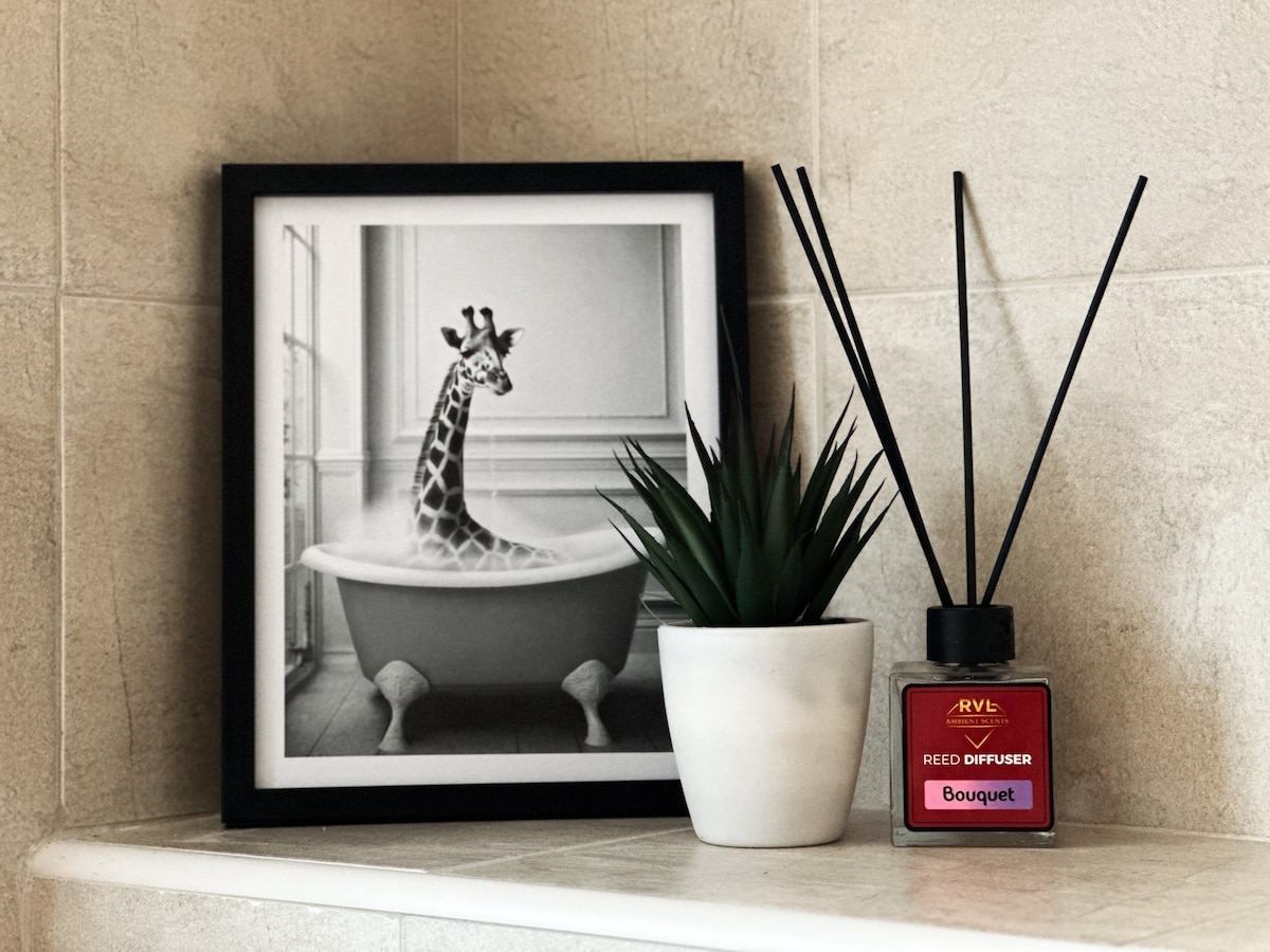 A framed black-and-white photograph of a giraffe in a bathtub rests on a countertop. Nearby, a small potted plant adds a touch of greenery, alongside a reed diffuser with a red label labeled 'Bouquet'. The minimalist arrangement contributes to a calming aesthetic.