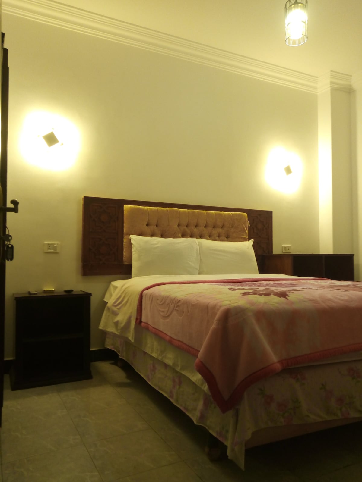 A well-lit bedroom features a spacious bed with a tufted headboard and crisp white linens. A soft pink blanket adds a cozy touch, with bedside tables on either side. Soft wall lights illuminate the room, enhancing a calm and restful ambiance.