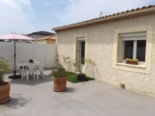 A light-colored building exterior is showcased, featuring a window adorned with potted plants. The outdoor area includes a white table and chairs under a pink parasol, surrounded by potted plants and a patch of green grass.