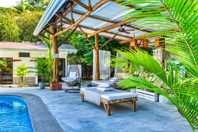 The outdoor lounge area features a spacious pool surrounded by tropical greenery. Comfortable day beds are arranged on a concrete deck, with soft cushions for relaxation. A hanging chair adds a unique touch, while a wooden roof provides shade over the seating area.