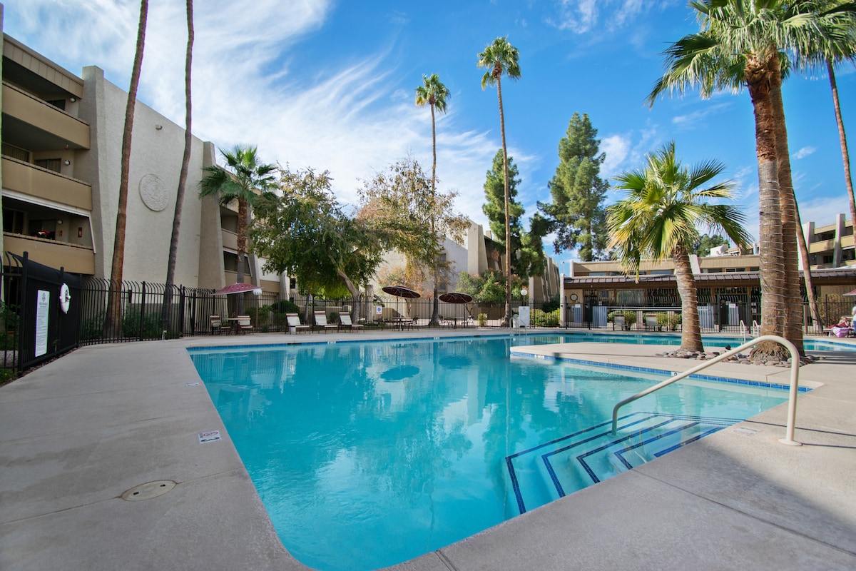 A resort-style swimming pool is featured, surrounded by palm trees and lounge chairs. The water appears clear and inviting, reflecting the blue sky above. Nearby, recreational areas and shaded seating options can be seen, enhancing the outdoor relaxation space.