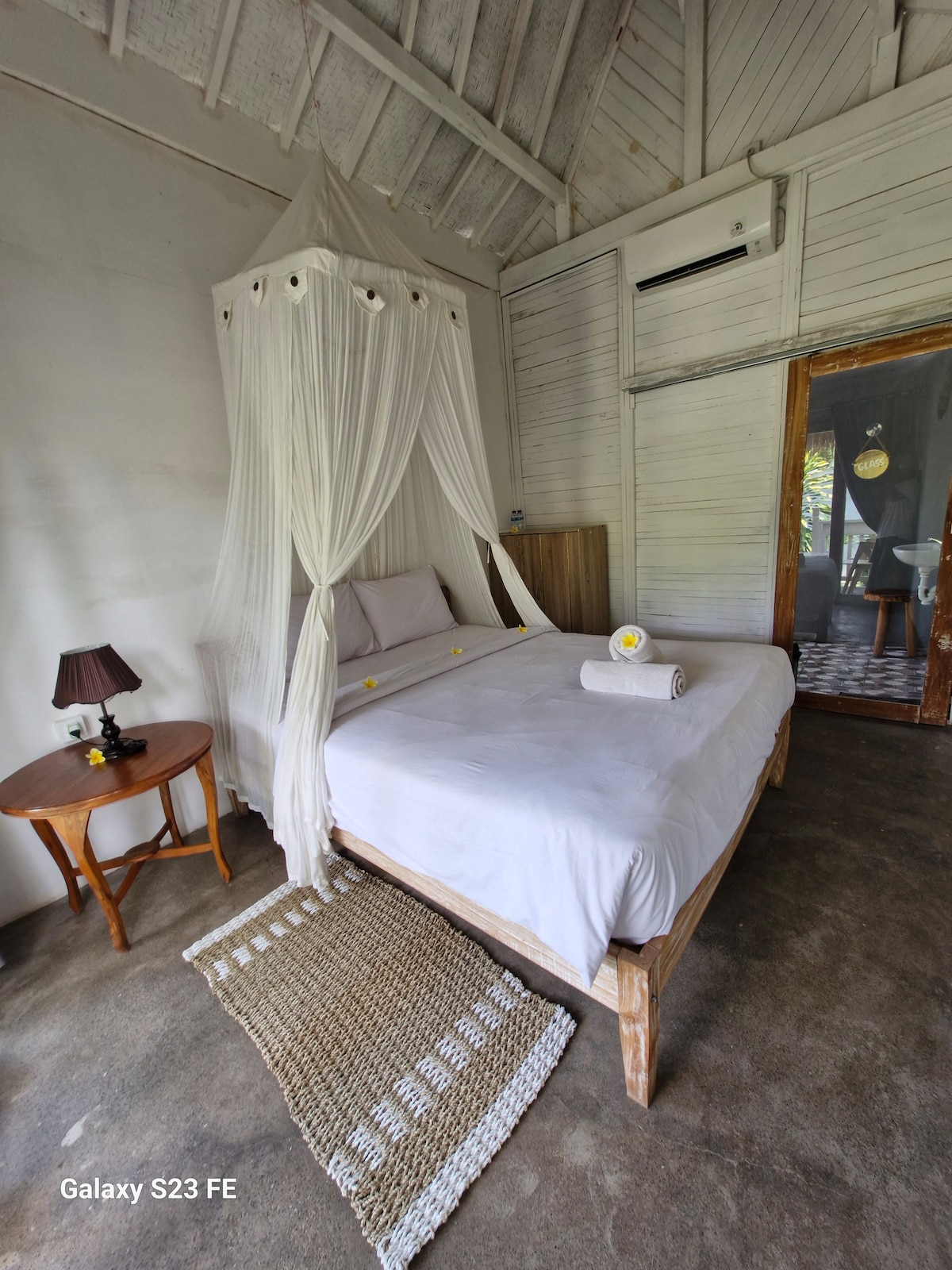 A spacious bed is draped with a sheer canopy, complemented by soft linens and decorative pillows. A wooden nightstand with a lamp sits beside the bed. Natural light filters through the room, enhancing the rustic wooden walls and concrete flooring.