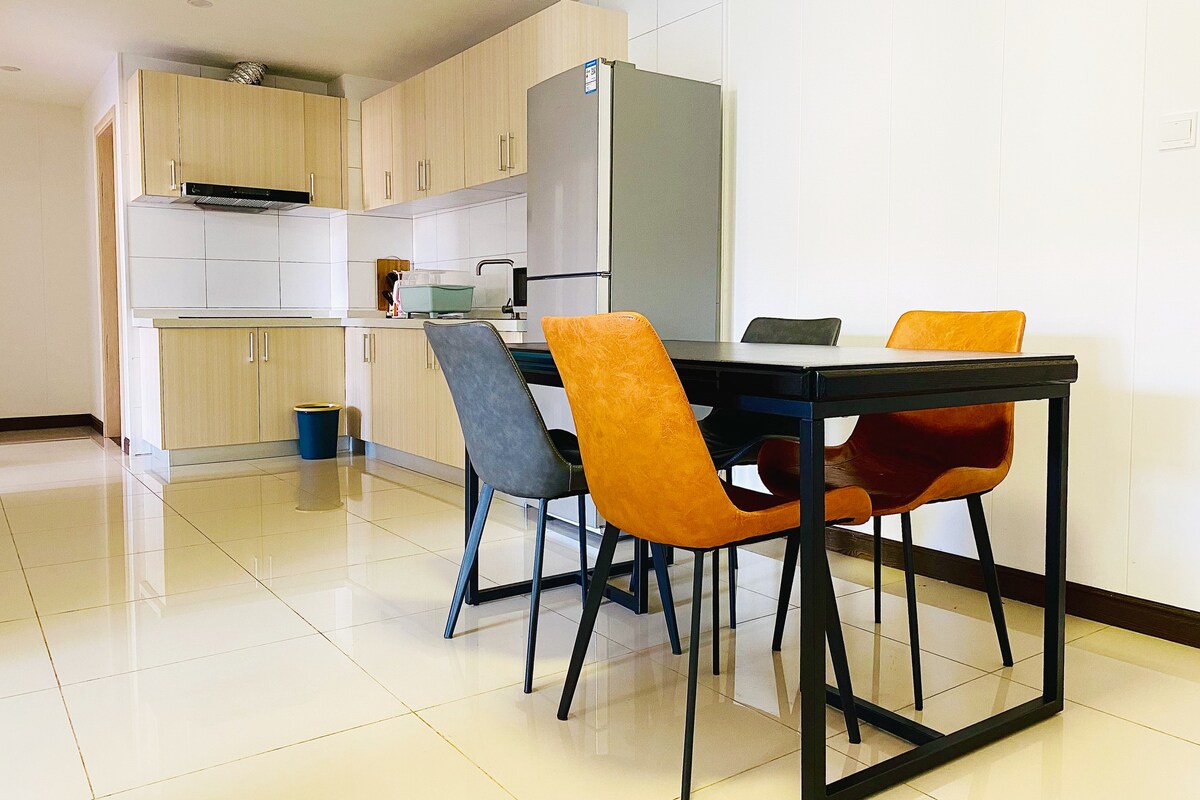 A modern dining area is presented with a sleek table surrounded by four chairs featuring orange and grey upholstery. The kitchen space is visible in the background, showcasing wooden cabinetry and a refrigerator. Bright tiles reflect available light, enhancing the open layout.