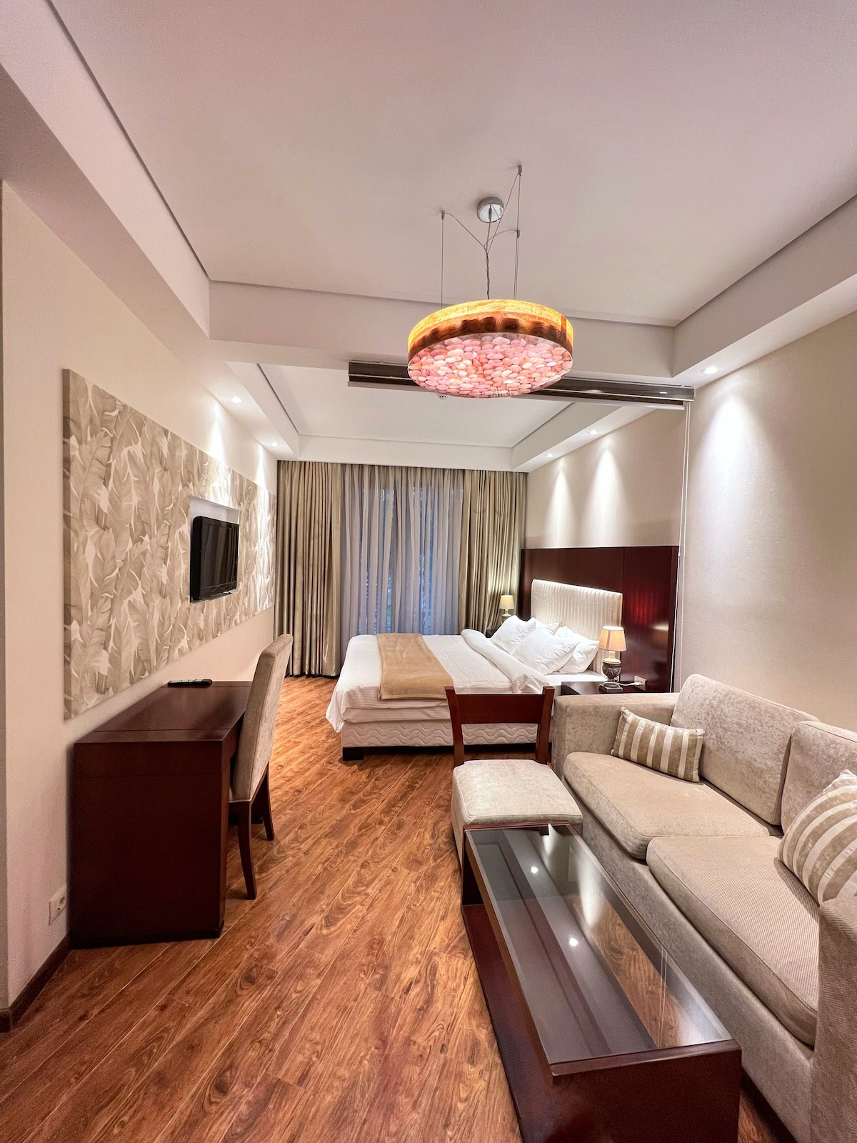 The studio features a king-size bed and a comfortable sofa with a coffee table. A wooden cabinet and a television are positioned against a wall. Soft lighting enhances the cozy ambiance, while large windows allow natural light to fill the space.