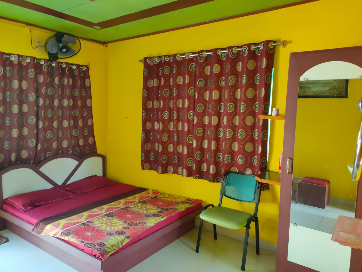 A non-AC room features bright yellow walls and contrasting maroon curtains decorated with a circular pattern. A spacious bed with a floral-patterned cover is positioned against the wall, accompanied by a simple chair and a mirror stand for convenience.