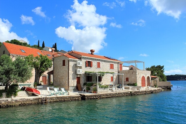 A stone building with red-tiled roofs is situated along the waterfront, surrounded by lush greenery. Lounge chairs are placed on a terrace facing the water, while the shimmering sea reflects the blue sky and clouds above, creating a serene atmosphere.