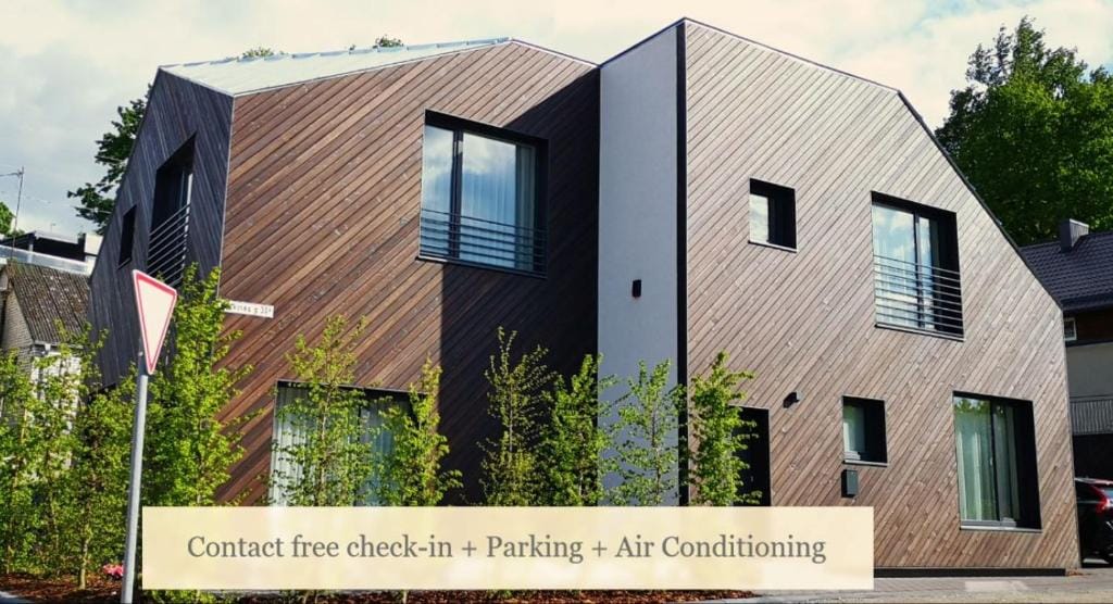 The exterior of the property features a distinctive larch wood design with modern angles. Large windows provide natural light, while surrounding greenery enhances the visual appeal. A sign noting amenities is displayed prominently, indicating contact-free check-in, parking, and air conditioning.