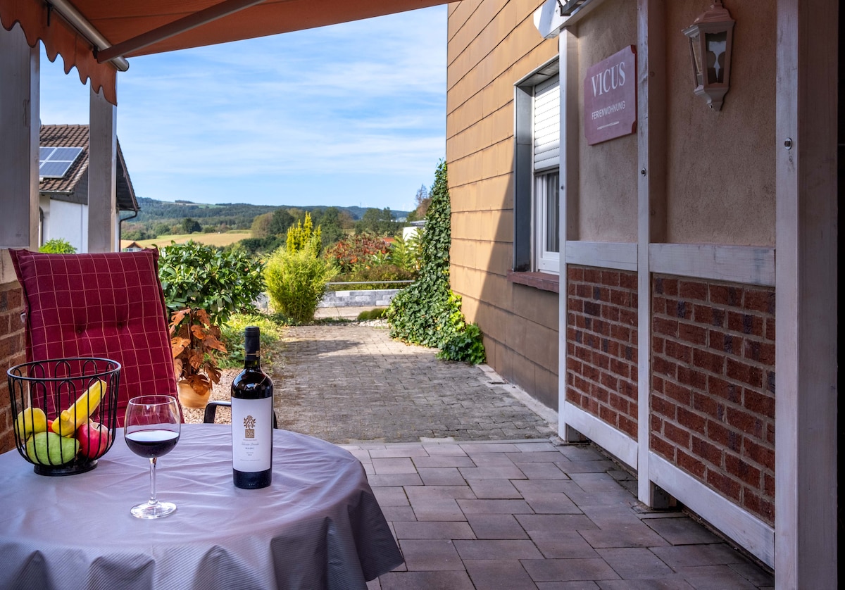An outdoor seating area is presented with a table covered in a light cloth. A bottle of wine and a glass rest on the table, accompanied by a basket of fresh fruit. The view features lush greenery and rolling hills in the distance.