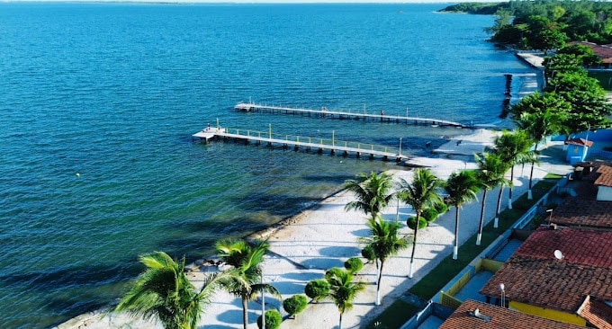The image captures a serene waterfront scene featuring a sandy beach lined with palm trees. A wooden dock extends into the calm blue water, while nearby structures with terracotta roofs are visible. The landscape is complemented by greenery, enhancing the tranquil environment.