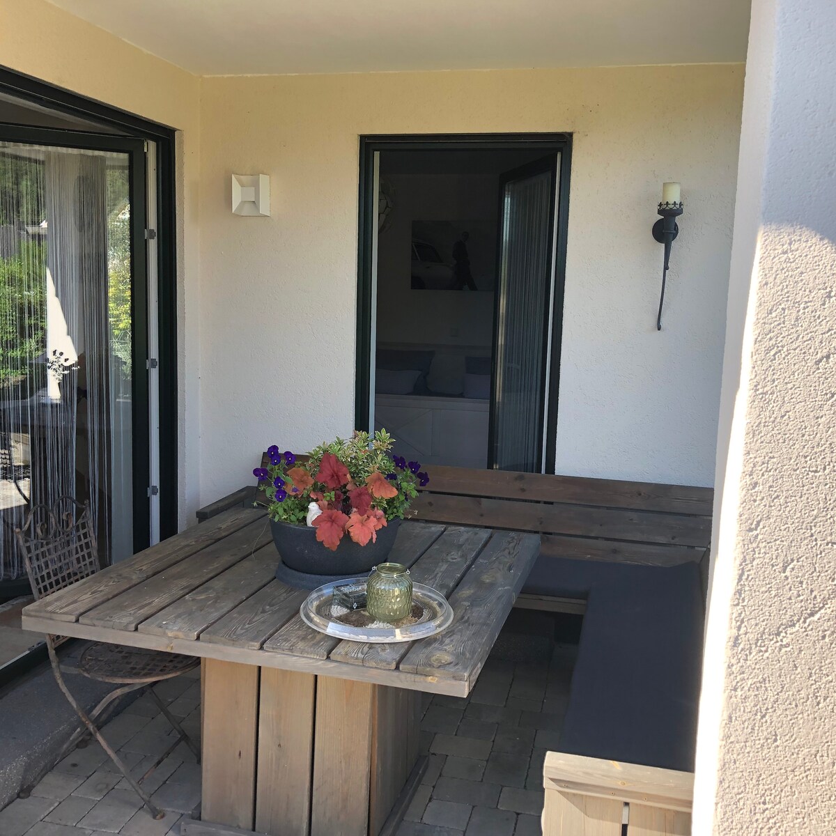 An outdoor terrace area features a rustic wooden table paired with built-in seating. A planter filled with vibrant flowers sits at the center, while an inviting atmosphere is created by the soft lighting fixtures mounted on the walls. Accessibility is provided through large glass doors.