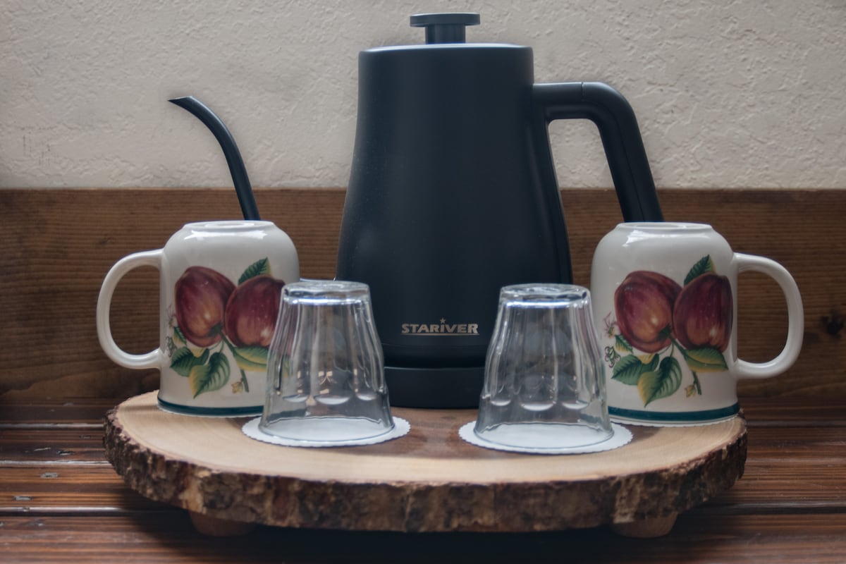 A stylish electric kettle is positioned at the center of a wooden serving tray. Two ceramic mugs, adorned with fruit illustrations, are placed beside two glass cups, all arranged neatly for easy access.
