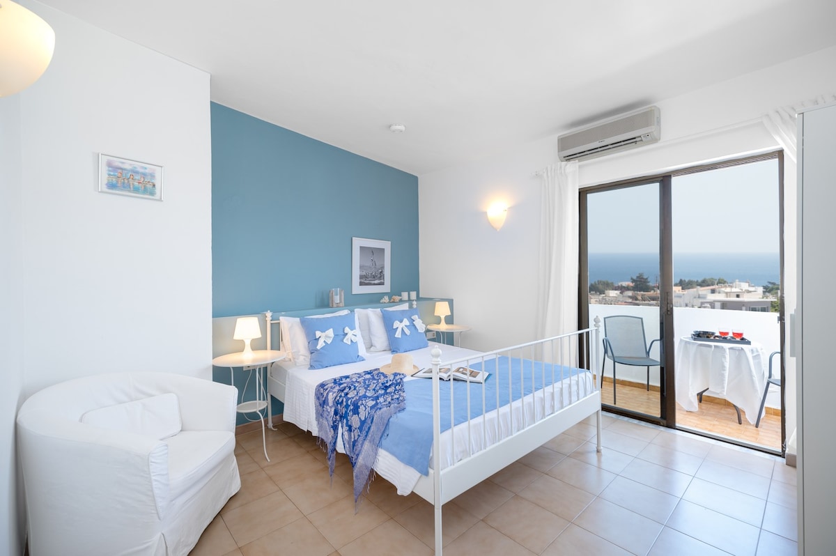 A spacious bedroom features a white metal bed dressed in light blue linens. Soft lighting is provided by bedside lamps. A cozy chair is placed in one corner, and a table with chairs is positioned on the balcony, offering sweeping views of the sea.