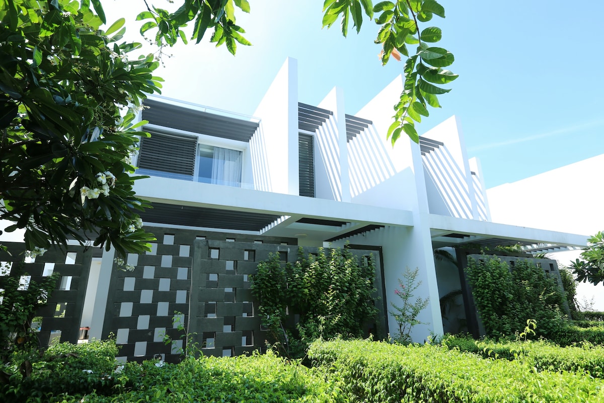 The villa's modern architecture is framed by lush greenery and vibrant foliage. The facade features a blend of horizontal elements and large windows, allowing natural light to filter through. The surrounding plants create a serene environment, enhancing the villa's peaceful ambiance.