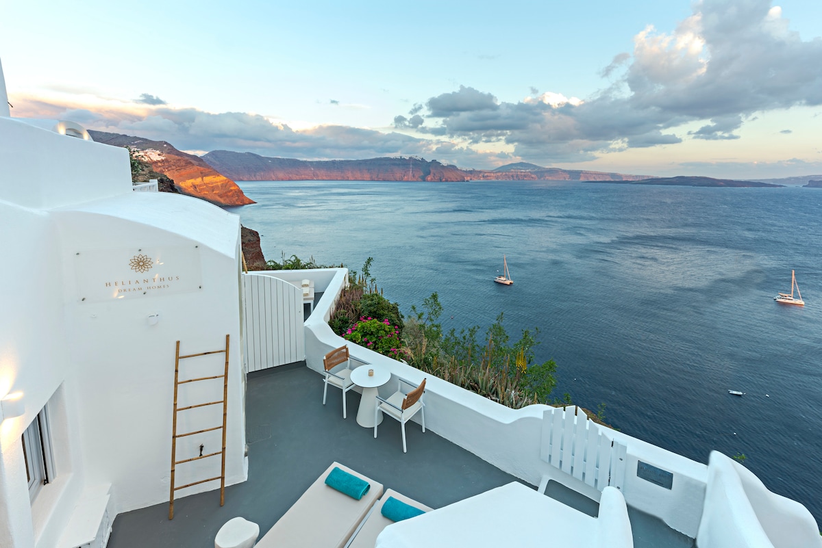 An outdoor terrace showcases views of the Aegean Sea and nearby coastlines. The space includes two lounge chairs, a small round table with two chairs, and delicate greenery. The horizon features sailboats and distant volcanic cliffs under a softly lit sky.