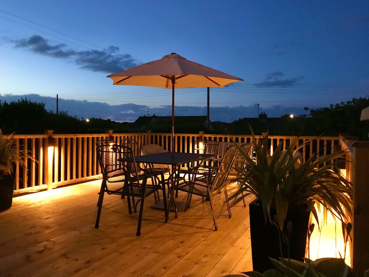 An outdoor deck area is illuminated by soft lighting, featuring a round dining table surrounded by chairs. A large umbrella provides shade. Potted plants are arranged around the space, creating an inviting setting for outdoor gatherings.
