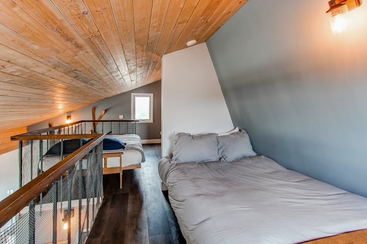 [Dhoma e gjumit 4] Krevati dopio "queen" dhe "futon" me madhësi të plotë qëndrojnë krah për krah në loft, me pamje nga mali nga dritaret. Pas kësaj fotografie ndodhet hapësira e punës, e përkryer për punë në distancë ose planifikimin e aventurës tënde të ardhshme.