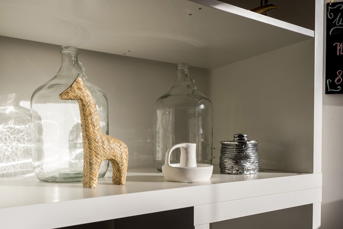 A minimalist shelf displays a collection of decorative items. A woven giraffe figurine stands alongside two clear glass bottles and a white sculptural vessel. A metallic spiral container adds texture and contrast, highlighted by soft, natural light.