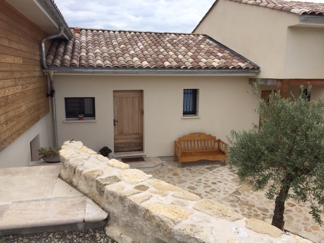 The exterior of the gîte features a rustic stone pathway leading to a wooden door. A wooden bench is positioned beside the entrance, surrounded by landscaped areas that include an olive tree and decorative plants. The roof showcases traditional tiles.