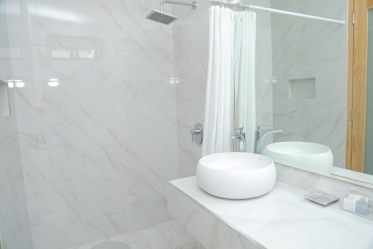 A modern bathroom is presented, featuring a spacious walk-in shower with a rainfall showerhead. A circular basin sits on a minimalist countertop, complemented by a large mirror. Light-colored marble tiles create a clean and elegant atmosphere.