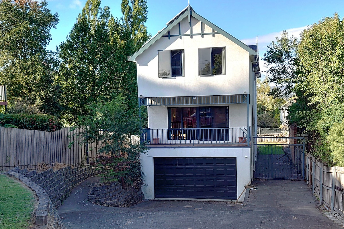 The exterior of a two-story home showcases a modern design with large windows and a balcony. A dark garage door is visible beneath the balcony, while a pathway leads to a secure yard surrounded by greenery. The building is set against a clear blue sky.