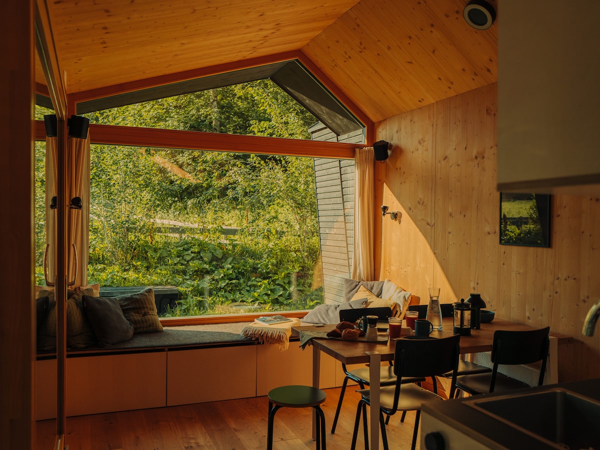 A cozy living area is highlighted by large windows that offer views of lush greenery. A dining table with chairs sits beside a comfortable seating nook adorned with cushions. Natural wood walls create a warm ambiance, illuminated by soft, natural light.