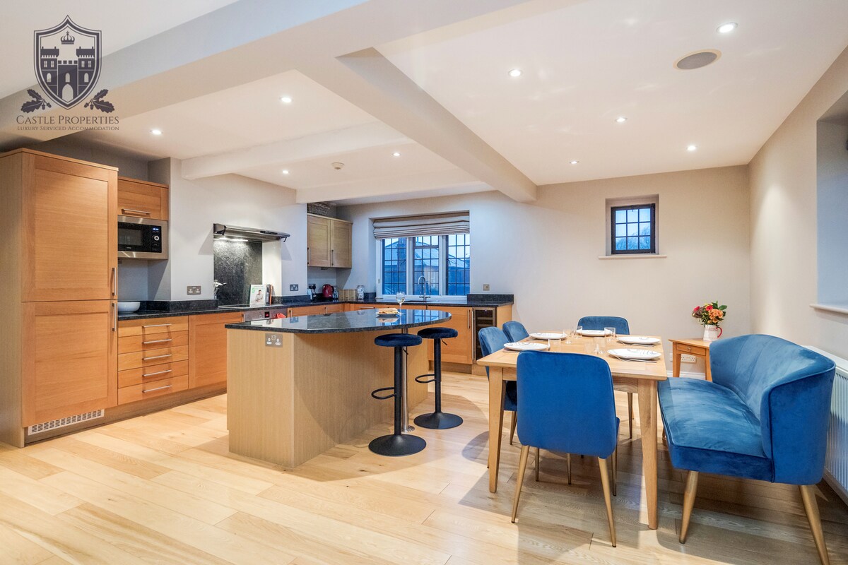 An open-plan kitchen and dining area features modern wooden cabinetry and a central island with bar stools. A wooden dining table is surrounded by blue upholstered chairs. Soft lighting enhances the warm tones of the wood flooring and walls, creating a welcoming space.
