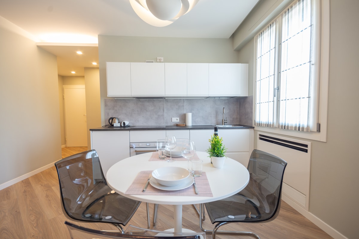 A modern kitchen area is highlighted by a round dining table set for two, featuring glassware and a small plant centerpiece. Sleek white cabinets and countertops complement the light wood flooring, while natural light fills the space through large windows with sheer curtains.