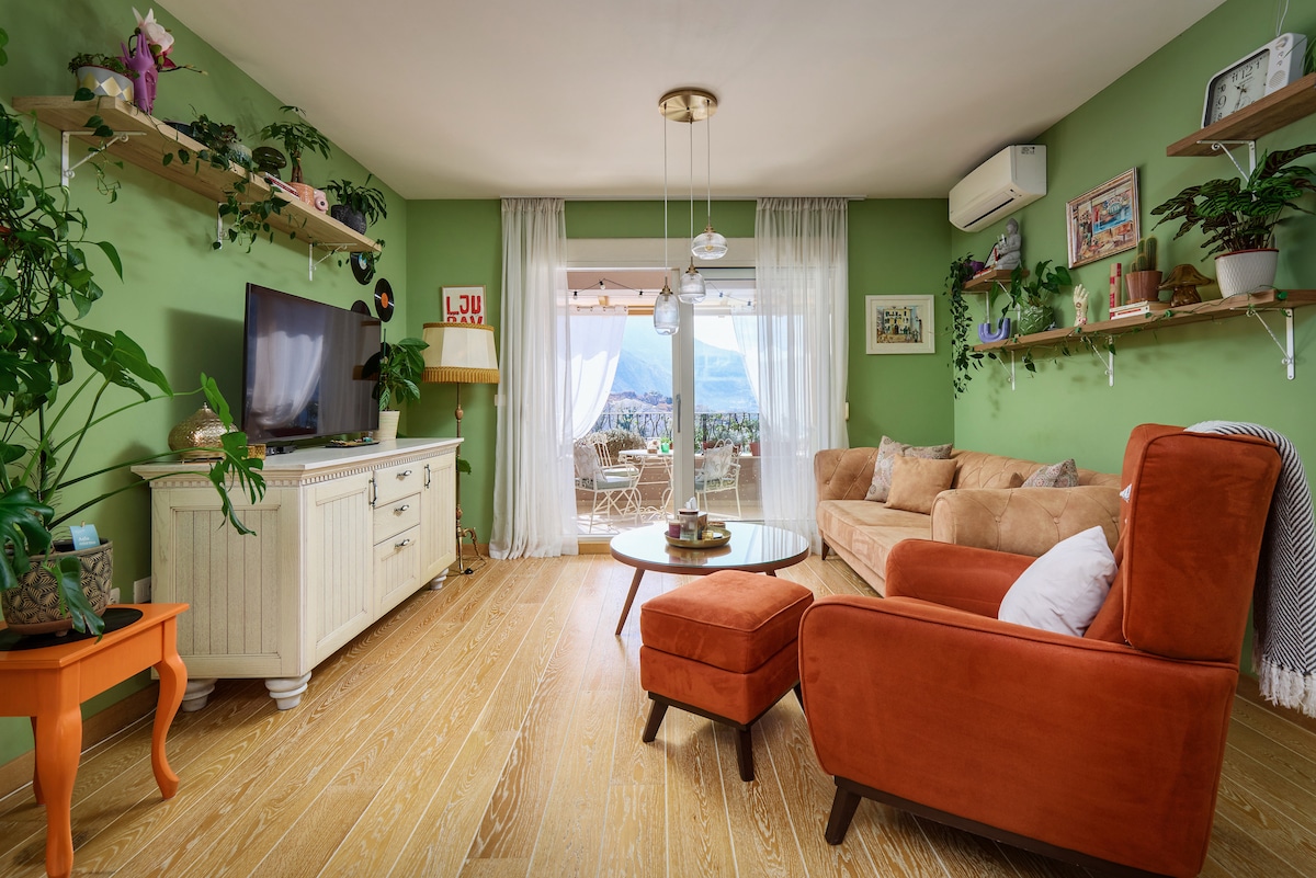 A cozy living area features green walls adorned with shelves holding plants and decor. A light wood floor is complemented by an orange armchair, a white TV console, and a round coffee table. Natural light enters through sheer curtains, revealing a view of the outdoors.
