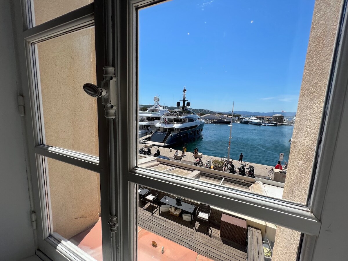 A view from a window showcases the bustling port of Saint Tropez. Large yachts are docked nearby, and the waterfront area is lined with outdoor seating, inviting a lively atmosphere. Bright blue skies complement the serene water, creating an open and airy feeling.