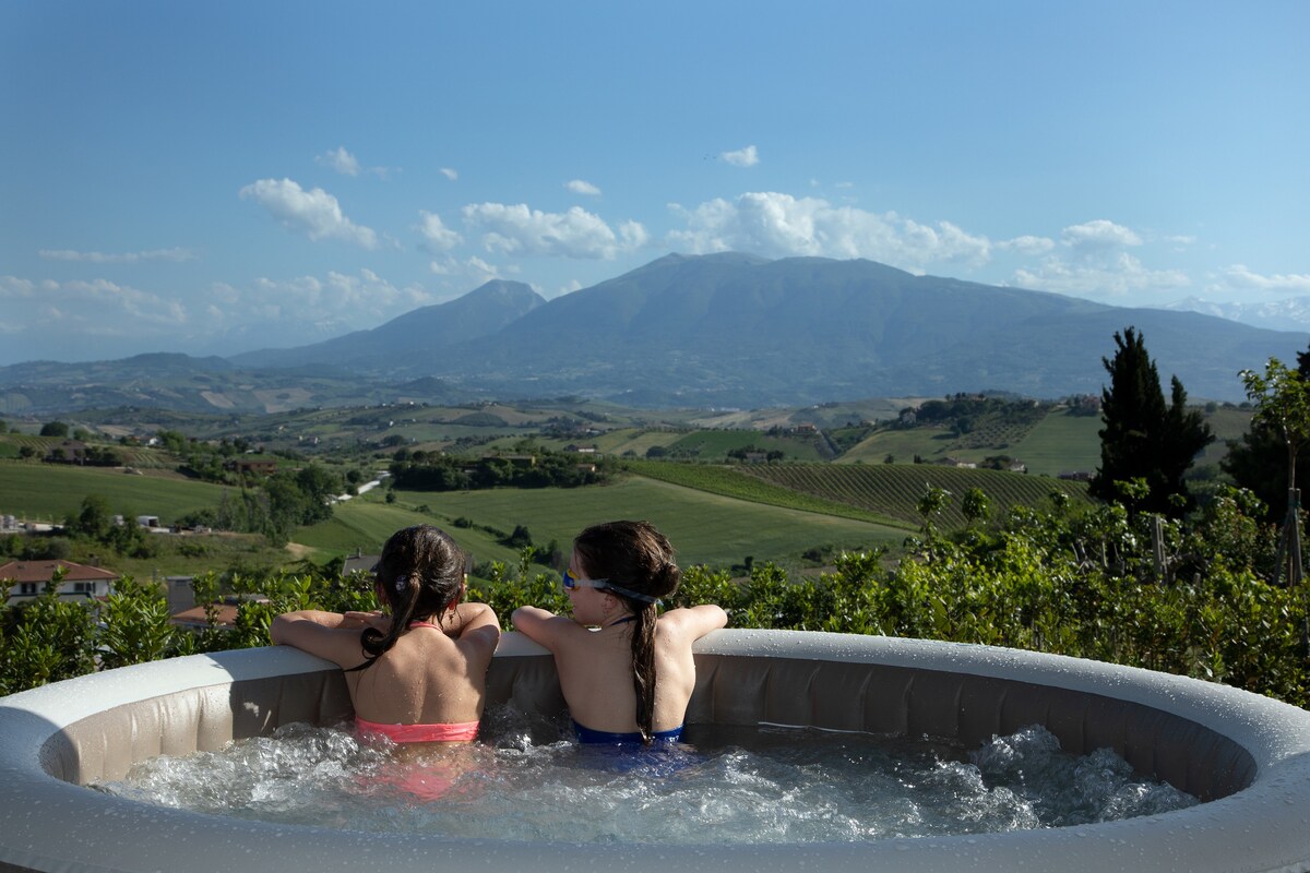 A hot tub is pictured on a terrace, offering an expansive view of rolling hills and distant mountains. Two children sit side by side, enjoying the water as gentle ripples create reflections accented by a clear blue sky.