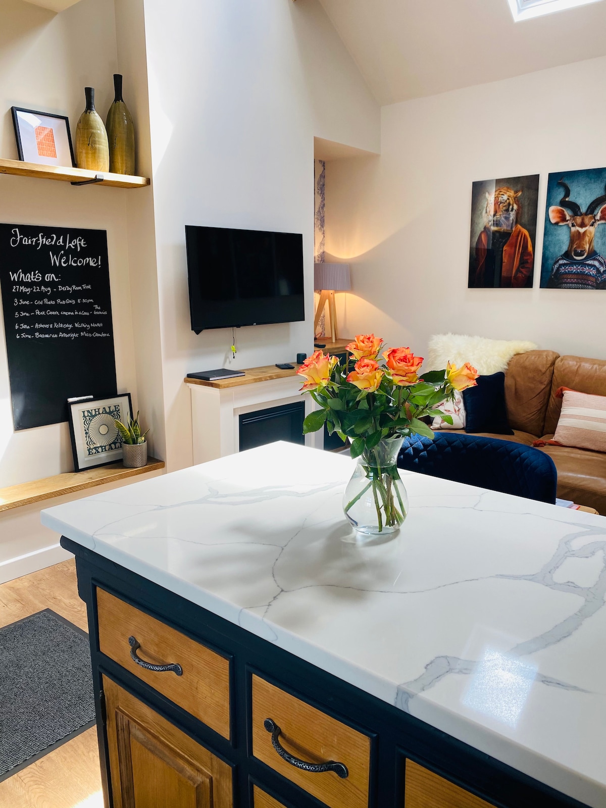 A modern open-plan living area features a well-equipped kitchen with a marble countertop and dark cabinetry. A vase with fresh roses sits on the counter. A comfortable sofa is observed in the background, alongside a decorative wall with framed artwork and a television.