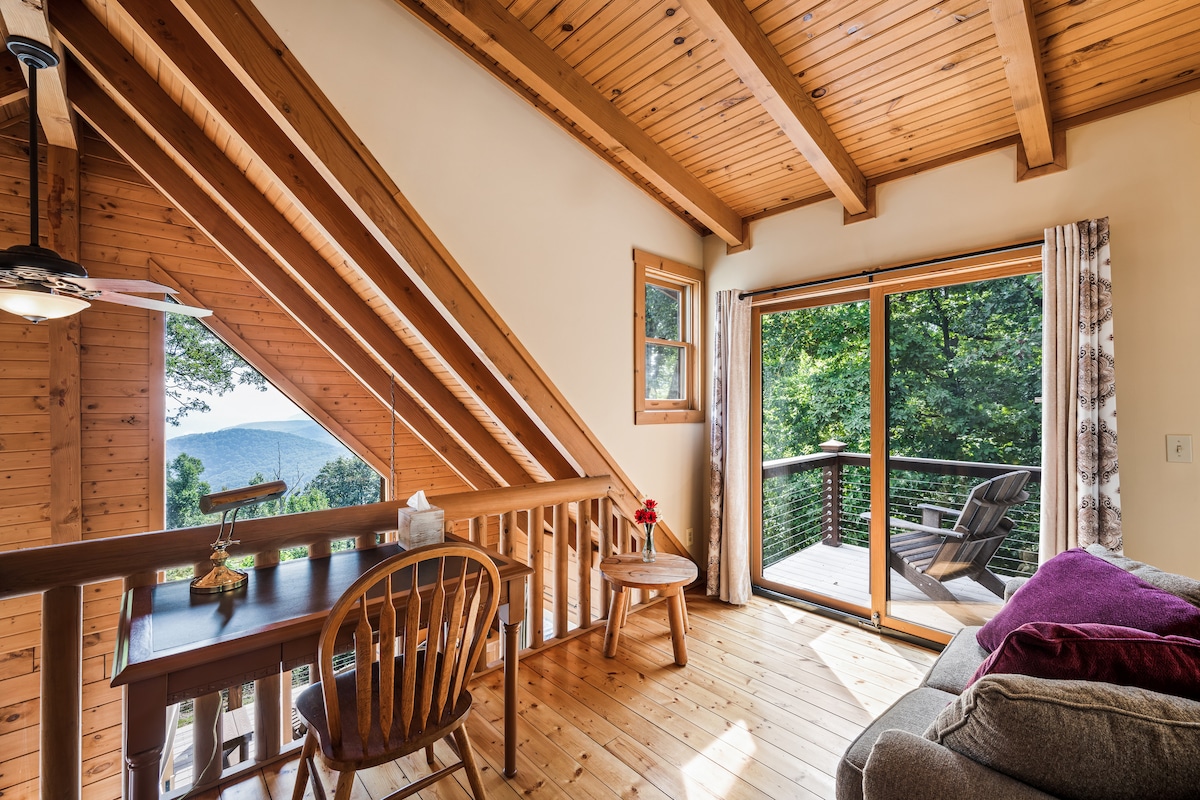 A cozy seating area is highlighted by natural wood beams and a large window with sliding doors that opens to a private deck. A small table with chairs is positioned near the window, creating a perfect spot for enjoying views of the surrounding trees and mountains.