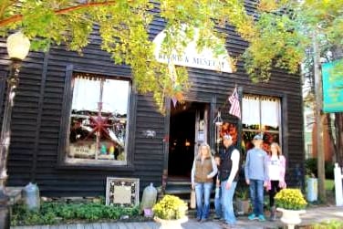 The entrance of a historic general store is framed by large windows with decorative curtains. A group of four individuals stands outside, engaged in conversation. Green bushes are positioned on either side of the entrance, and flags add a festive touch to the scene.
