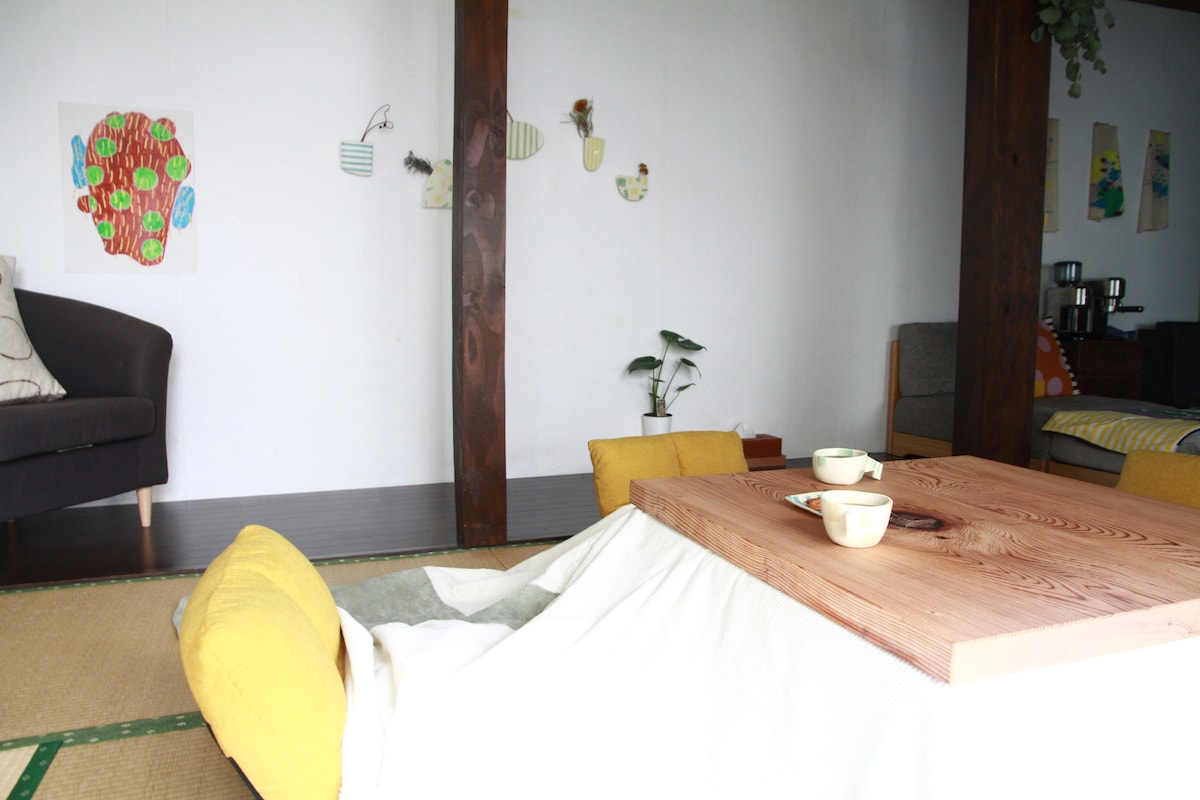 A well-arranged living area features a low wooden table surrounded by yellow cushions. Walls are adorned with colorful artworks, and a small indoor plant adds a touch of greenery. A cozy armchair is positioned in a corner, contributing to the inviting atmosphere.