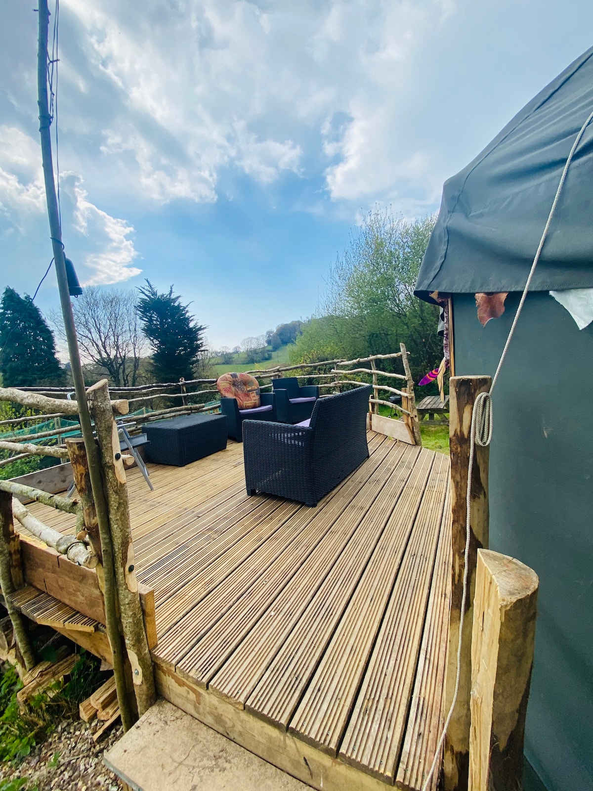 A wooden terrace is highlighted, featuring two comfortable patio chairs. The space is surrounded by a natural landscape with trees visible in the background, under a partly cloudy sky. The terrace offers an inviting area to relax and enjoy the outdoors.