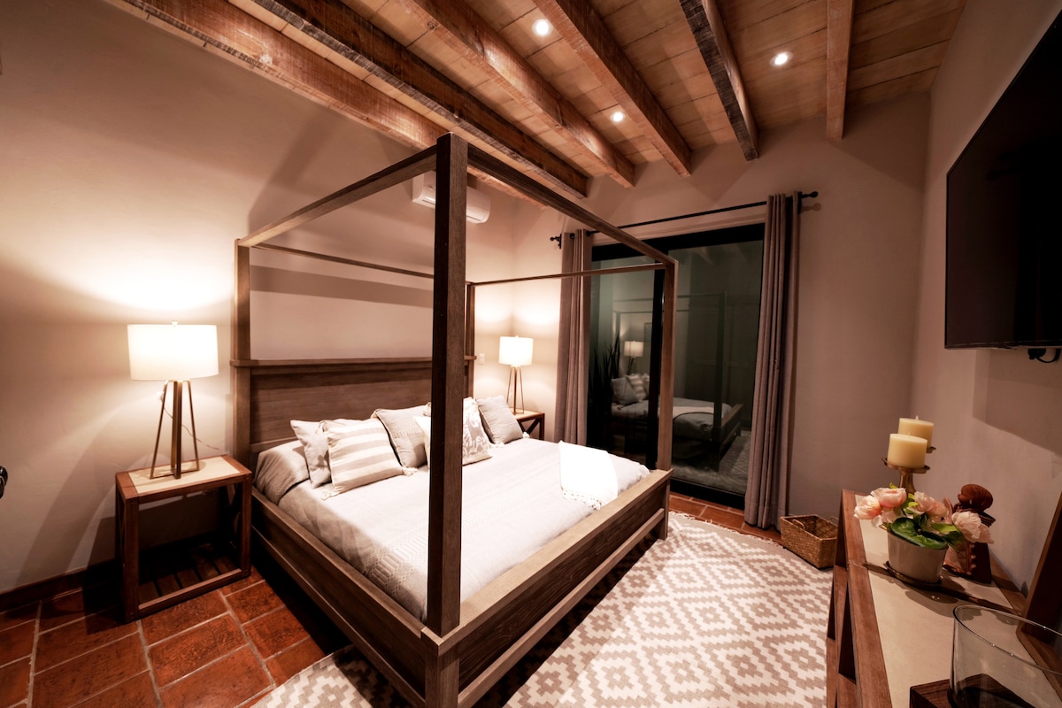 A spacious bedroom features a four-poster bed adorned with light-colored bedding. Soft lighting from bedside lamps enhances the welcoming environment. A large window draped in dark curtains provides additional light, while a textured area rug and wooden furniture add warmth to the space.