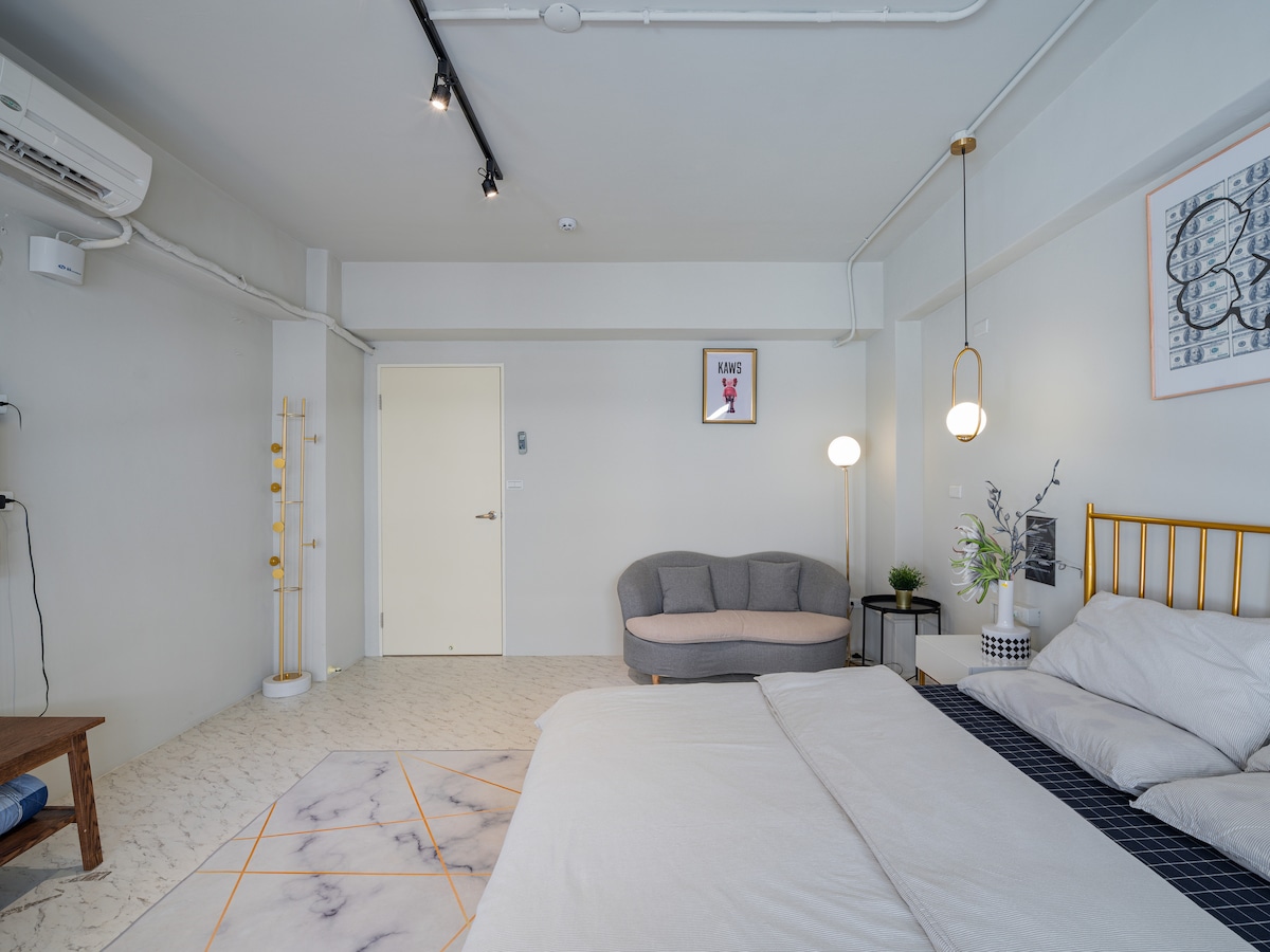 A spacious bedroom is featured, with a large bed and soft bedding inviting relaxation. A gray sofa is positioned nearby, complemented by stylish lighting and minimalist decor. The room includes light-colored walls and a contemporary design, creating an airy and comfortable atmosphere.