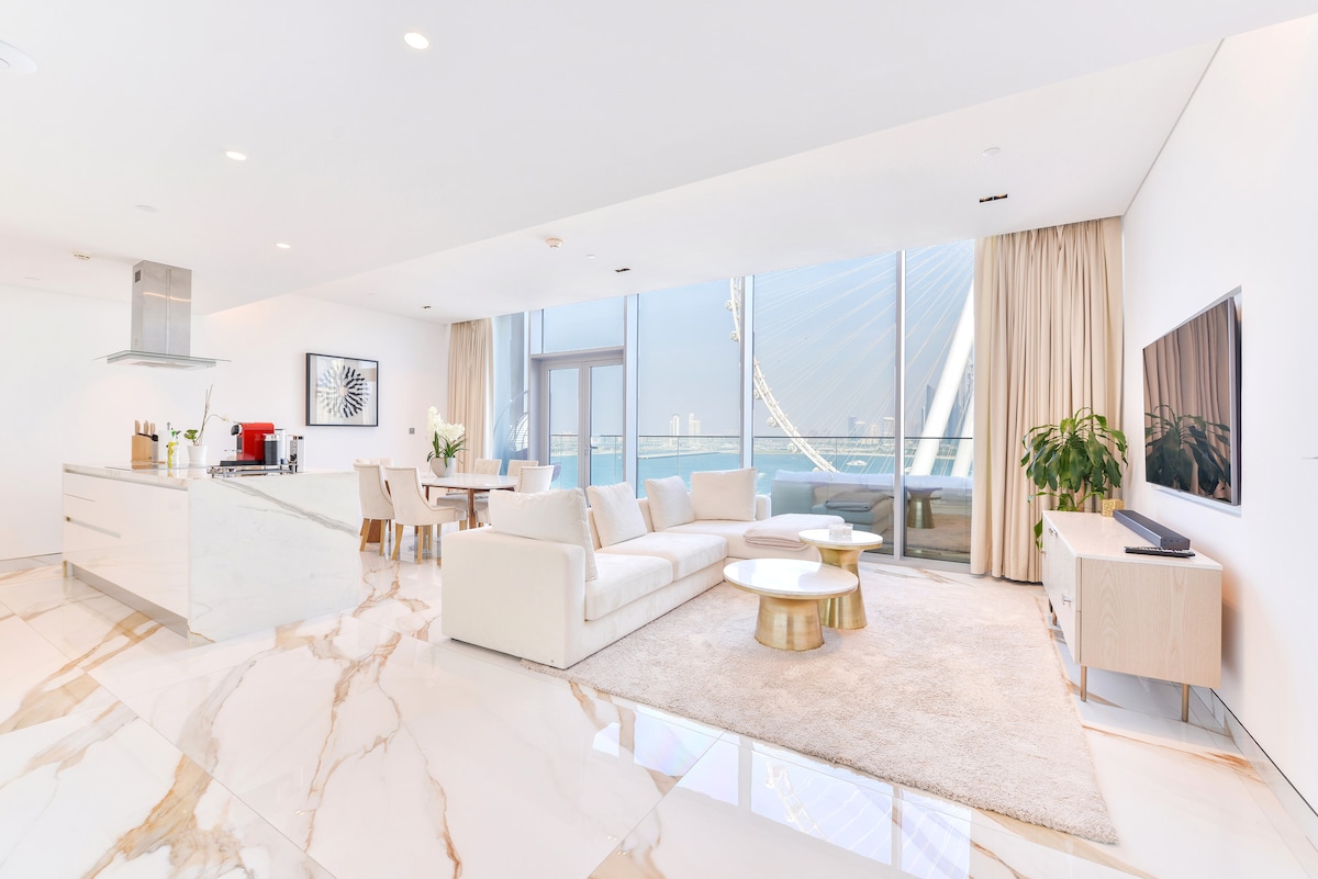 The modern living area showcases a spacious layout with a light-colored sectional sofa and a minimalist coffee table. Large windows provide ample natural light and stunning views of Ain Dubai. A sleek kitchen area with a marble countertop complements the elegant decor.