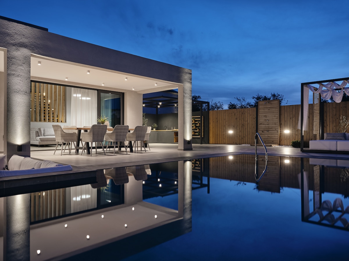 An elegant outdoor dining area is visible, featuring a large table surrounded by several chairs. The ambient lighting reflects beautifully in the calm pool water, enhancing the serene atmosphere as the sun sets, highlighting the villa's modern architecture.