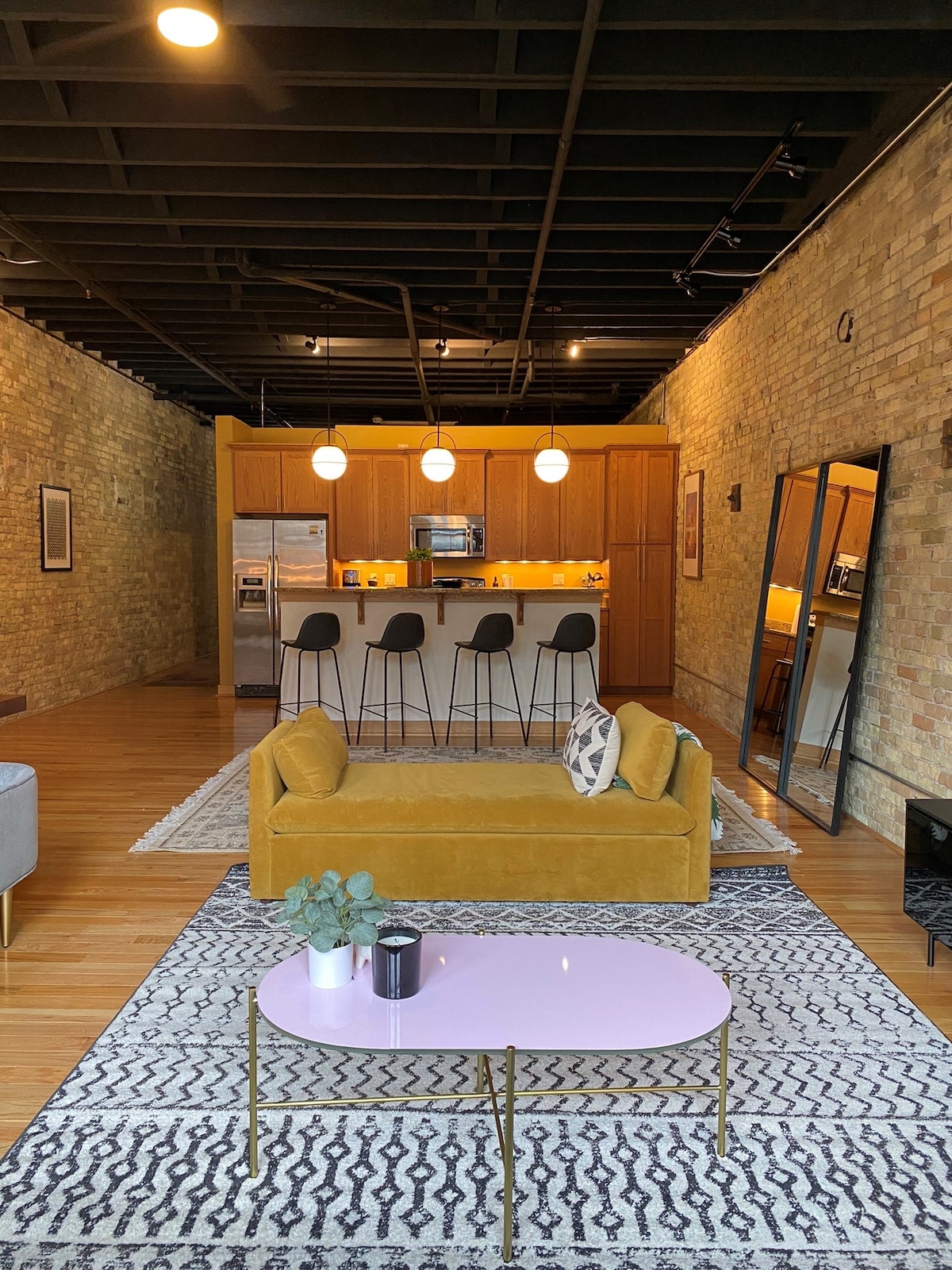 The spacious living area features cream city brick walls and polished hardwood floors. A yellow couch and patterned rug create a comfortable seating arrangement. A pink coffee table is centered, while a well-equipped kitchen with modern appliances is visible in the background, illuminated by pendant lighting.