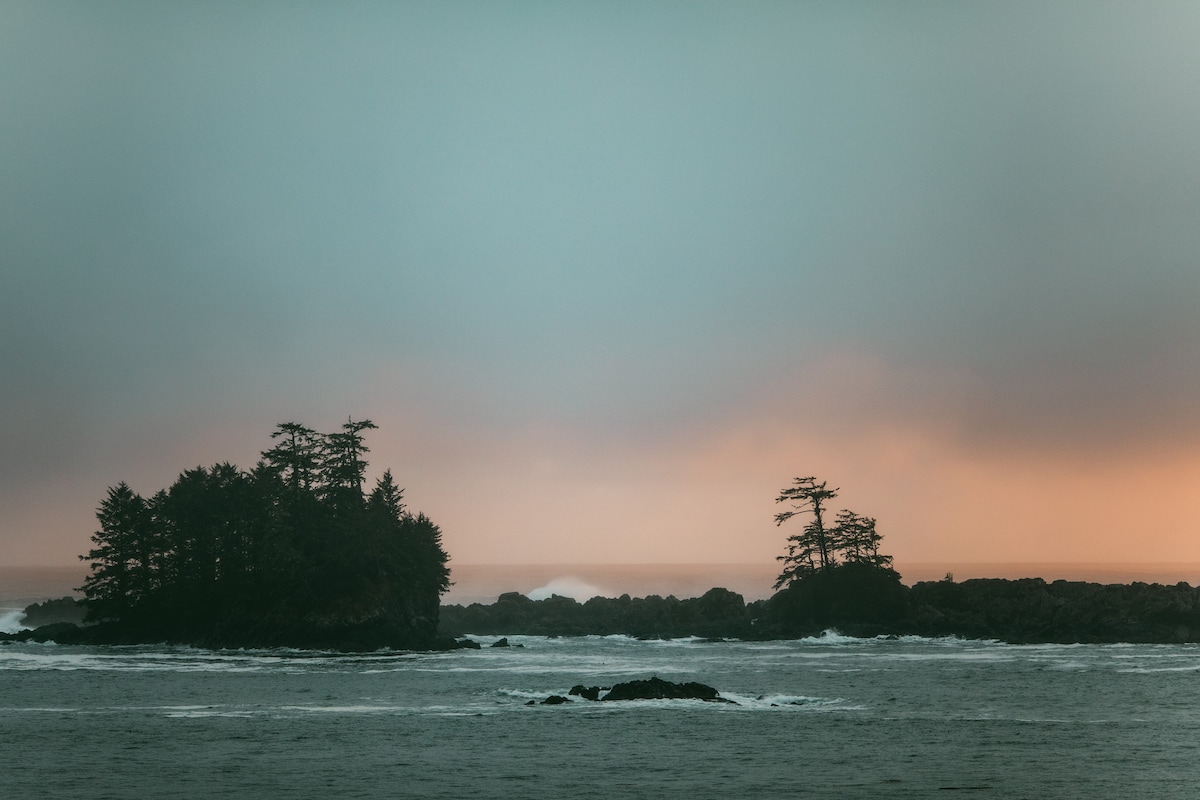 Two rugged islands emerge from the ocean, surrounded by gentle waves and a misty atmosphere. Dark green trees populate the islands, contrasting with a soft gradient of orange and gray in the sky. A tranquil scene captures the essence of the coastal landscape.